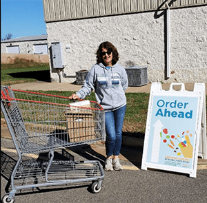 A volunteer stands by an Order Ahead sign with a shopping cart.