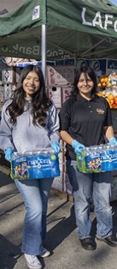 Two young women holding cases of water in front of a tent.