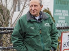 An older man leans against a fence outside a soup kitchen.