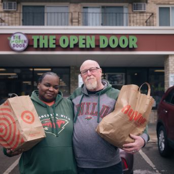 a couple holding paper grocery bags stand in front of The Open Door Pantry