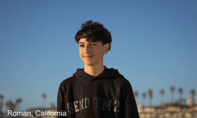 Roman stands on a beach in California wearing a black hoodie, looking out toward the ocean under a clear blue sky.
