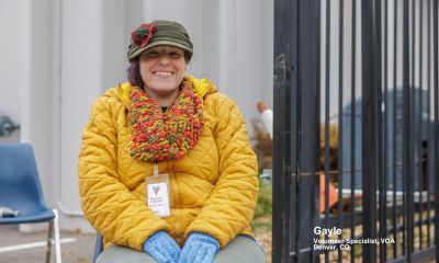 A smiling Gayle wearing a yellow quilted jacket, a colorful scarf, and an olive cap sits outdoors in Denver, CO, as a VOA Volunteer Specialist.