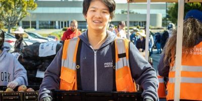  A teenager wearing an orange vest smiles while holding a box filled with fresh, red apples.
