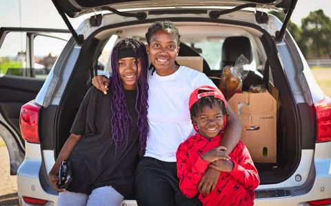 A mom and her two children smiling in front of their car.