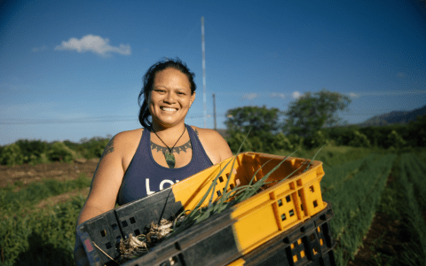 A smiling Hawaiian woman holds a crate of fresh vegetables