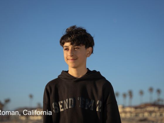Roman stands on a beach in California wearing a black hoodie, looking out toward the ocean under a clear blue sky.