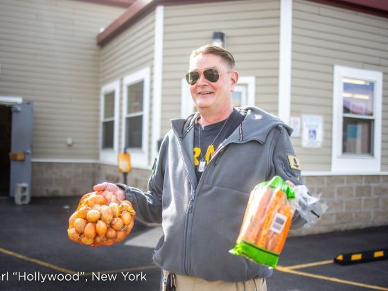 Carl "Hollywood" smiles outside a food distribution site in New York while holding bags of onions and carrots.
