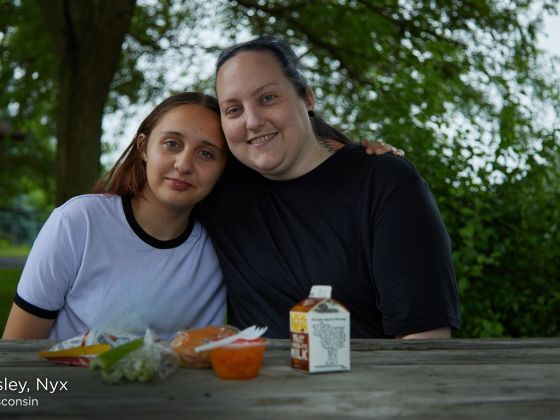 Nyx and her daughter Eisley sit together at a picnic table outdoors in Wisconsin, smiling with their arms around each other. Fresh fruit, a carton of milk and other snacks sit on the table in front of them.