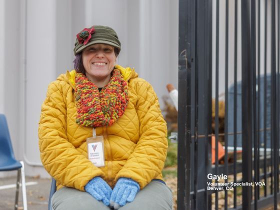 A smiling Gayle wearing a yellow quilted jacket, a colorful scarf, and an olive cap sits outdoors in Denver, CO, as a VOA Volunteer Specialist.