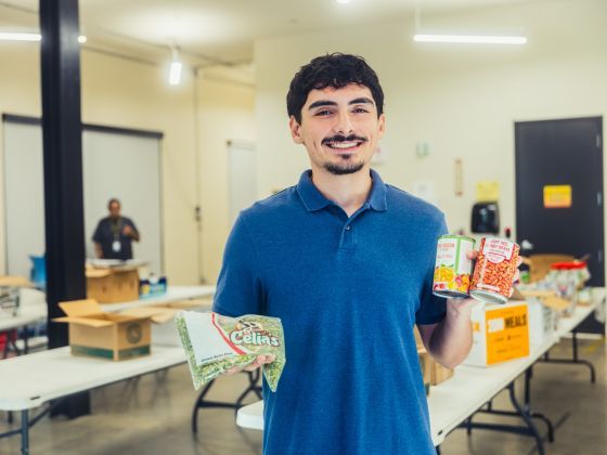 A smiling Angel wearing a blue polo shirt holding various non-perishable food items