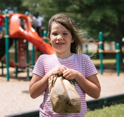 A child holding a lunch bag in front of a park.