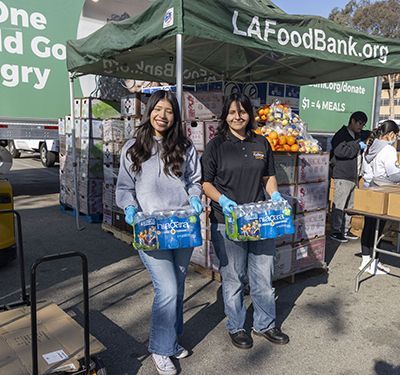 Two young women holding cases of water in front of a tent.