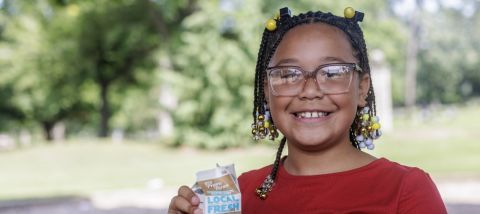 A smiling young girl with glasses drinking chocolate milk outside.
