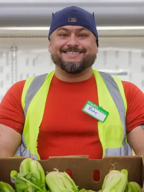 Food bank volunteer holding box of lettuce in front of shelf of produce.