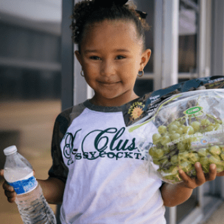 Young girl holding plastic water bottle and a bag of grapes.