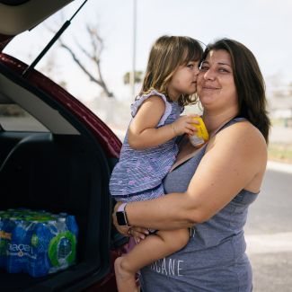 A mom holding her daughter in front of the opened trunk of her car.
