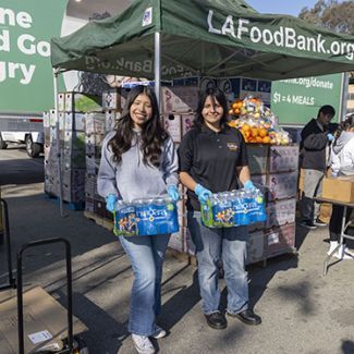 Two young women holding cases of water in front of a tent.