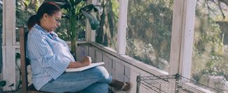Woman sitting on porch writing in notebook, leg crossed over knee.