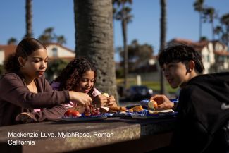 Roman and his sisters, Mackenzie-Luve and MylahRose, sit together at an outdoor table in California eating snacks and fruit on a sunny day.
