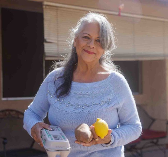 A woman in a blue sweater holds a carton of eggs, a sweet potato, and a lemon, standing outdoors.