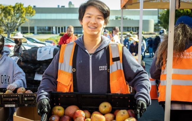  A teenager wearing an orange vest smiles while holding a box filled with fresh, red apples.