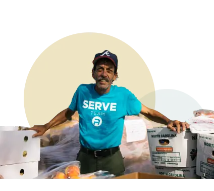 Smiling older gentleman with a t-shirt saying 'Serve' standing on front of cases of produce